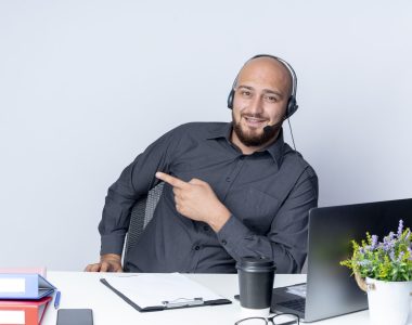 impressed young bald call center man wearing headset sitting at desk with work tools pointing at side isolated on white background