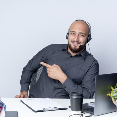 impressed young bald call center man wearing headset sitting at desk with work tools pointing at side isolated on white background impressed young bald call center man wearing headset sitting at desk with work tools pointing at side isolated on white background
